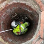 Birds eye perspective of a man working in a deep, round manhole. He wears bright yellow PPE and a white hard hat, and is attached securely to a white rope that leads out of the manhole.