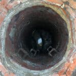 Top-down perspective of run-down manhole before works. The manhole is open, round and is similar shape to a well, with dirty, old red bricks forming the walls of the manhole. Debris such as mud and sewerage is building up in the base.