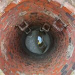 Top-down perspective of a deep, round manhole being filled, from the base, with a light-grey grout. There is a pool of water at the base, revealing the reflection of the man, in bright PPE, taking the photo.