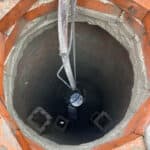 Birds-eye perspective of a deep, round manhole. A piece of equipment called a spinner is attached to a hose and is being fed into the manhole. It's job is to coat the inner walls of the manhole with grout and seal the manhole from infiltration and exfiltration.
